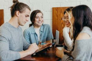 Four young adults sit around a wooden table, smiling and talking. One person uses a tablet, discussing web site design ideas for their marketing agency as the group enjoys a casual, friendly conversation in a relaxed setting.