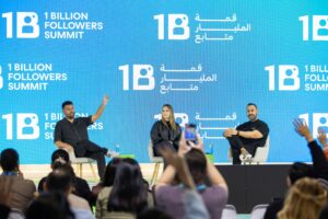 Three speakers sit on stage at the 1 Billion Followers Summit, engaging with an audience. Two men and one woman discuss social media management in front of a blue backdrop with summit branding, while attendees eagerly raise their hands.