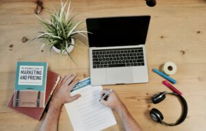 A person writes on a paper at a wooden desk with an open laptop, a book titled Marketing and Pricing, plant, headphones, sticky notes, tape, ruler, blue marker—perfect for brainstorming web site design or digital marketing ideas.