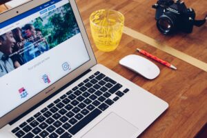 A wooden desk with an open MacBook displaying a Facebook ads page, perfect for digital marketing tasks, alongside a white Apple Magic Mouse, red pencil, yellow glass filled with paperclips, and a black camera.