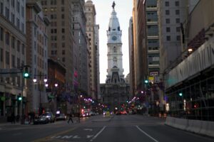 A view down a city street in Philadelphia with traffic and pedestrians, leading to the historic City Hall, is perfect inspiration for a marketing agency specializing in web site design and social media management amid tall buildings at dusk.