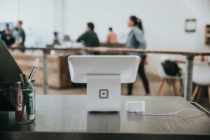 A white Square point-of-sale terminal sits on a countertop in a modern cafe, with people conversing in the background. A cup with pens, a card reader, and materials from a marketing agency are also visible on the counter.