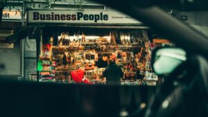 Two people stand in front of a busy street newsstand called Business People, filled with magazines and newspapers—a classic spot that contrasts the fast pace of digital marketing outside, viewed through a car window for a blurred foreground effect.