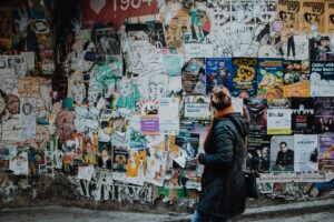 A person in a dark coat and orange scarf walks past a wall covered with colorful posters, flyers, and graffiti art—reflecting the vibrant creativity often found in digital marketing and social media management.