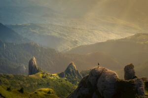 A person stands on a rocky peak surrounded by lush green hills and mountains, with soft sunlight streaming through a hazy sky—capturing the kind of tranquil, majestic landscape perfect for a digital marketing campaign.