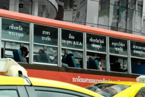 A red and orange city bus with Thai writing on its windows is filled with seated passengers. Yellow taxis are in the foreground, while city buildings—perhaps home to a digital marketing agency or web site design firm—are reflected in the bus windows.