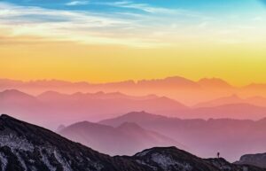 A vibrant sunset casts orange, pink, and purple hues over layers of distant mountain ranges, with a lone person standing on a rocky ridge—an inspiring scene perfect for a marketing agency’s web site design or digital marketing campaign.