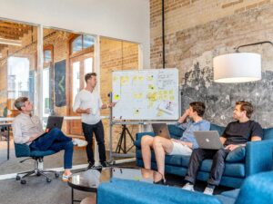 Four men in a casual marketing agency setting discuss at a whiteboard covered with sticky notes and diagrams. Three are seated with laptops, while one stands and leads the meeting focused on web site design strategies.
