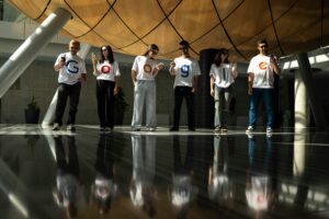Six people stand indoors, each wearing a white shirt with a single letter; together, their shirts spell Google. As they look at their phones, reflecting on the shiny floor, they appear ready to brainstorm digital marketing or web site design ideas.