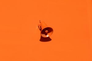 A wall-mounted loudspeaker painted entirely in orange casts a shadow on a plain orange wall, creating a minimalist and monochromatic composition reminiscent of modern marketing agency aesthetics.