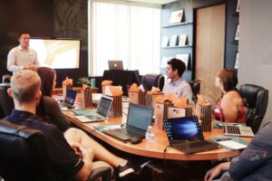 A man presents to five seated colleagues in a conference room. Laptops, water bottles, and striped gift bags are on the table. A large screen displays a Social Media Management slide as daylight streams through vertical blinds.