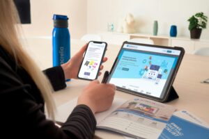 A person sits at a desk using a smartphone and tablet, both displaying digital marketing platforms. A blue water bottle, notebook, pen, and marketing agency brochure are also on the light-colored desk.