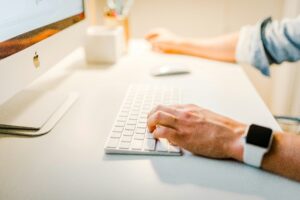 A person wearing a smartwatch types on a white keyboard at a desk with a computer monitor and office supplies, working on web site design in a bright workspace.