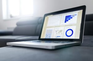 A laptop on a dark sofa displays charts and graphs—line graphs, bar graphs, a world map, and a blue circular chart—suggesting data analysis or business analytics for digital marketing or social media management.