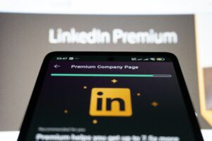 A smartphone screen displays the LinkedIn Premium Company Page, highlighting social media management tools, with the LinkedIn logo visible. In the blurred background, the words LinkedIn Premium appear on a monitor.