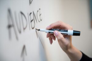 A hand holding a black dry-erase marker writes the word “AUDIENCE” in large letters on a whiteboard, illustrating key concepts in digital marketing and web site design.