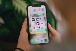 A person holds a smartphone displaying a folder of social media apps—a perfect scene for any marketing agency or digital marketing campaign—with a leafy background blurred behind.