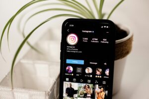 A smartphone displays the Instagram profile page, highlighting digital marketing success with 7,065 posts and 473 million followers. The phone sits before a green potted plant and white textured container on a light surface.