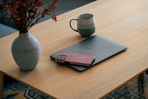 A closed laptop, a smartphone with a pink patterned case, and a ceramic mug sit on a light wooden table next to a vase with dried branches—an inspiring setup for creative social media management.