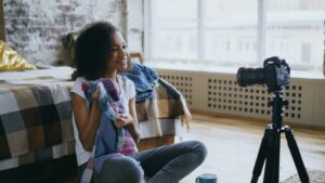 A woman sitting on the floor in a cozy bedroom holds up clothes and smiles at a camera on a tripod, possibly recording a vlog or fashion video about digital marketing. Natural light fills the room through large windows.