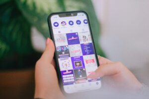 A person holds a smartphone with both hands, scrolling through a vibrant social media profile managed by a marketing agency. Multiple posts and icons are visible on the screen, with green leaves blurred in the background.
