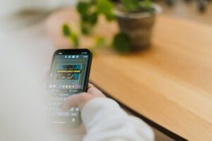 Person holding a smartphone, composing a message with a “DOWNLOAD HERE” image on the screen—perfect for digital marketing or social media management. A potted plant and wooden table are visible in the blurred background.