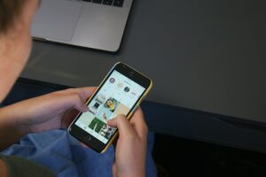 A person uses a smartphone to browse an Instagram profile, with several photos visible on the screen, while sitting at a desk with a laptop partially visible—perfect inspiration for social media management or marketing agency tasks.
