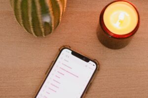 A smartphone displaying a list of hashtag searches lies on a wooden surface next to a lit candle in a brown holder and a green cactus in a pot—perfect inspiration for social media management or digital marketing creativity.