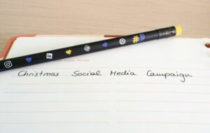 A pencil decorated with social media icons rests on an open notebook. The page is labeled Christmas Social Media Campaign, showcasing a marketing agency’s creative approach to digital marketing in neat handwriting.