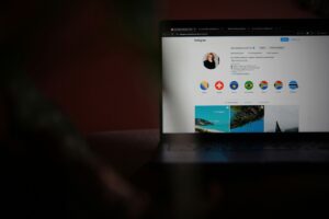 A laptop displaying an Instagram profile with story highlights like flag icons and travel photos is open in a dimly lit room. The scene hints at social media management, with the screen partially visible amid the dark surroundings.
