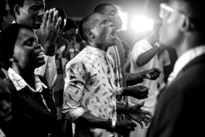 A group of people, mostly men, stand closely together indoors. One man in the center passionately shouts or sings, eyes closed, hands clenched. The mood is intense and emotional; it could be a team at a marketing agency celebrating a web site design win. Bright lights shine behind them.