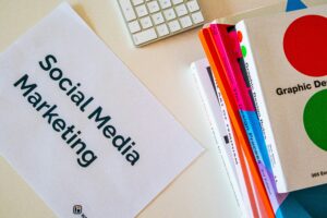 A paper labeled Social Media Marketing lies on a desk next to colorful books, including one titled Graphic Design, highlighting the essentials of digital marketing, with a partial view of a computer keyboard nearby.