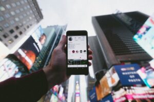 A person holds up a smartphone displaying an Instagram profile, with tall city buildings and bright billboards in the blurry background, highlighting the role of social media management in a vibrant urban setting like Times Square.