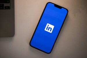 A smartphone displaying the LinkedIn logo on a blue background sits on a light-colored surface next to the edge of a laptop, highlighting tools essential for social media management and web site design.