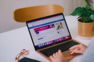 A person manages social media on a rose gold laptop displaying Facebook. Nearby, orange glasses, a black notebook, and a potted plant rest on a white table—perfect for digital marketing or web site design tasks.