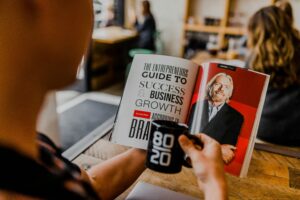 A person holds a black “80/20” mug while reading a magazine in a café, featuring “The Entrepreneurs Guide to Success & Business Growth”—perfect inspiration for anyone exploring web site design or working with a marketing agency.