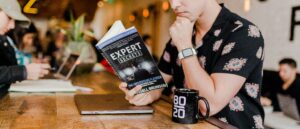 A person in a floral shirt reads Expert Secrets by Russell Brunson at a wooden table with a laptop and a black 80/20 mug, in a busy café—a perfect setting for inspiration in digital marketing or social media management.