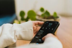 A person wearing a white sleeve holds a smartphone, interacting with a social media management app, while a plant in a pot sits blurred in the background on a wooden surface.