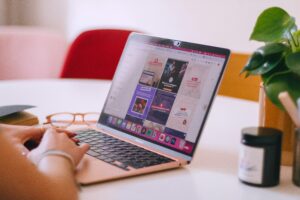 A person types on a rose-gold laptop displaying a digital marketing project. Glasses, a notebook, a candle, and a potted plant are on the white table. In the background sits a red chair, suggesting work for a marketing agency.