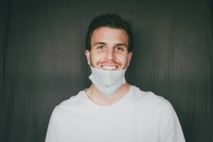 A young man wearing a white shirt and a face mask pulled down below his chin smiles while standing in front of a plain dark background, ready to discuss web site design or share digital marketing insights.