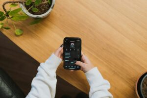 A person in a white sweatshirt holds a smartphone, viewing a music app, at a wooden table with potted plants nearby—perfect inspiration for web site design or social media management by a marketing agency.