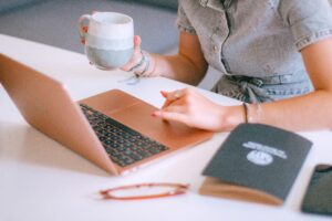 A person in a short-sleeve dress uses a rose gold laptop while holding a ceramic mug. Nearby are a notebook, eyeglasses, and a pen on a white desk—perfect for brainstorming digital marketing or web site design ideas.