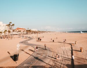 A wide, sandy beach with a paved bike and walking path is alive with people biking, walking, and relaxing. Palm trees and buildings line the background under a blue sky. The ocean glistens on the right—a perfect spot for a marketing agency’s website design shoot.