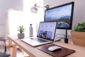 A tidy, modern desk setup with a laptop, monitor, mousepad, stylus, water bottle, small potted plants, and a wall lamp. The monitor displays “WORK HARD ANYWHERE” above a scenic landscape.