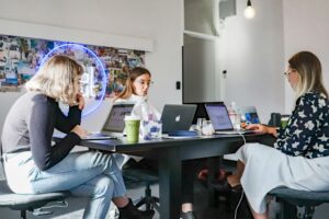 Three women sit around a table in a modern office, working on laptops. Coffee cups and papers are on the table, and a neon sign and collage of photos decorate the wall in the background.