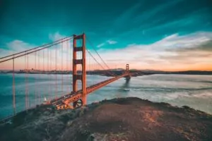 The Golden Gate Bridge stretches across the water at sunset, with a partly cloudy sky above and a boat passing beneath the bridge. The city skyline is visible in the background.